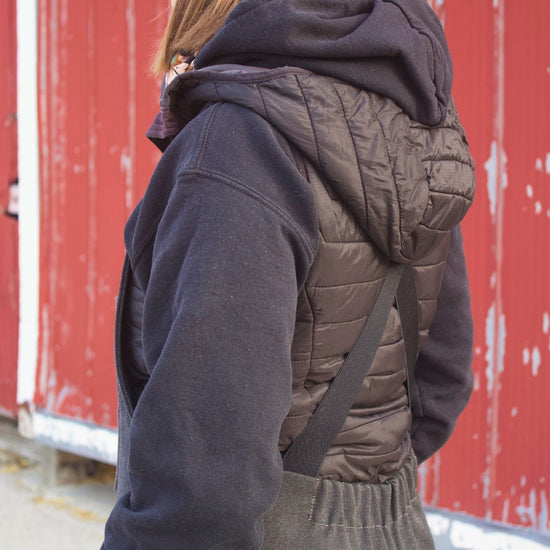 Person wearing a dark jacket, gray vest, and pink beanie in front of a red barn.