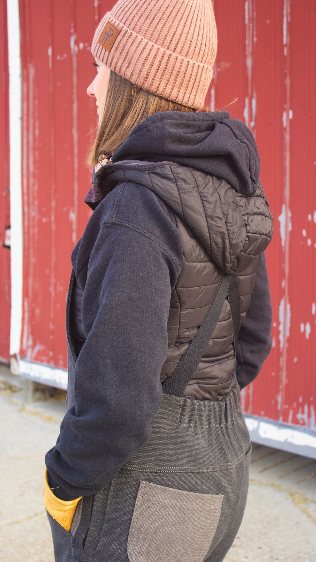 Person wearing a dark jacket, gray vest, and pink beanie in front of a red barn.