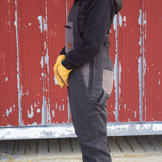 Person wearing a black jacket, grey overalls, yellow gloves, and a brown beanie standing in front of a red barn.