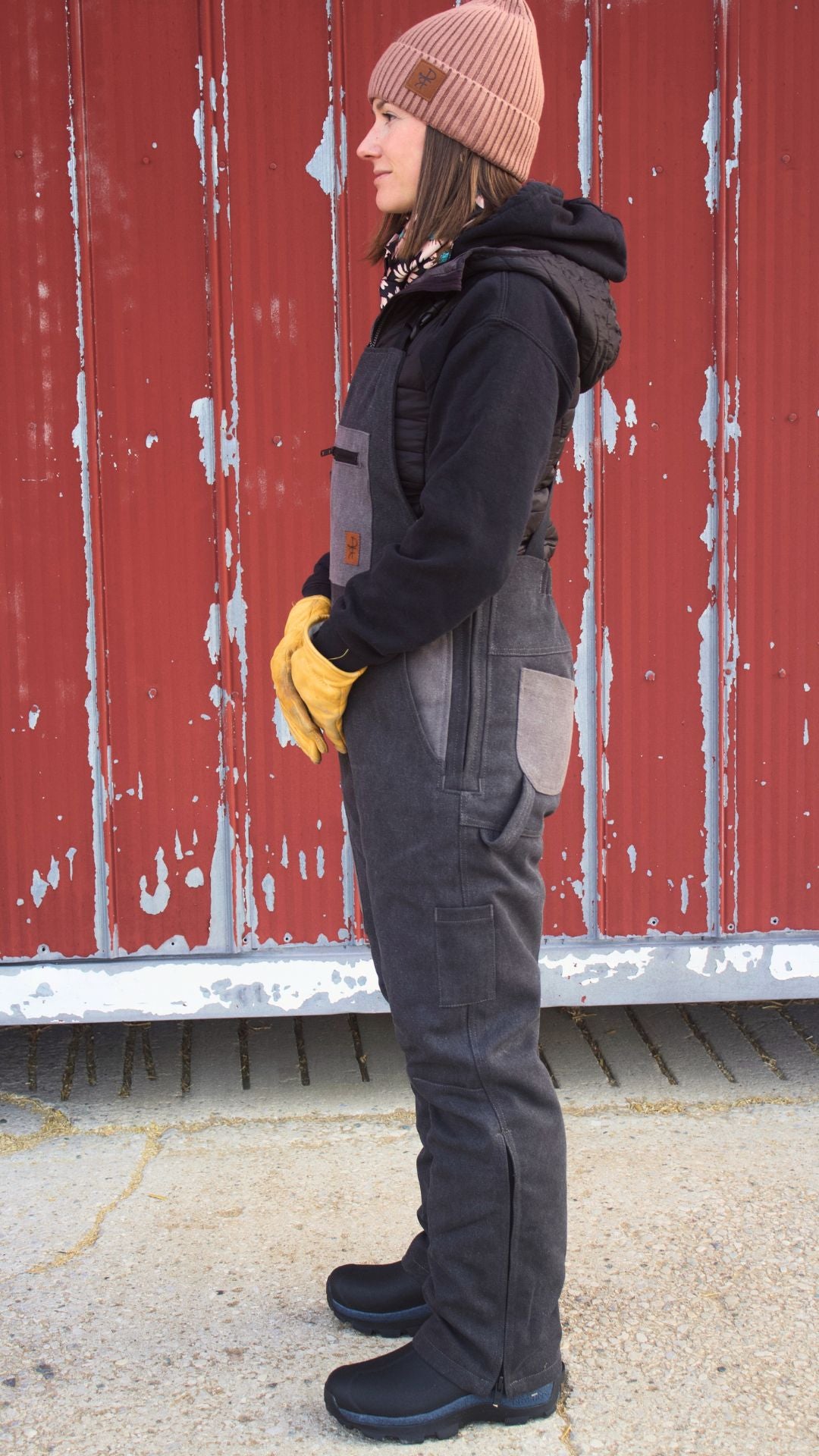 Person wearing a black jacket, grey overalls, yellow gloves, and a brown beanie standing in front of a red barn.