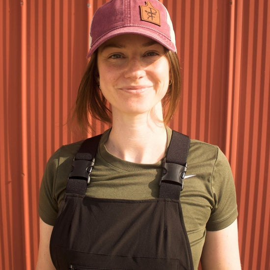 Person wearing a green shirt, black overalls, and a maroon cap against a red metal wall.