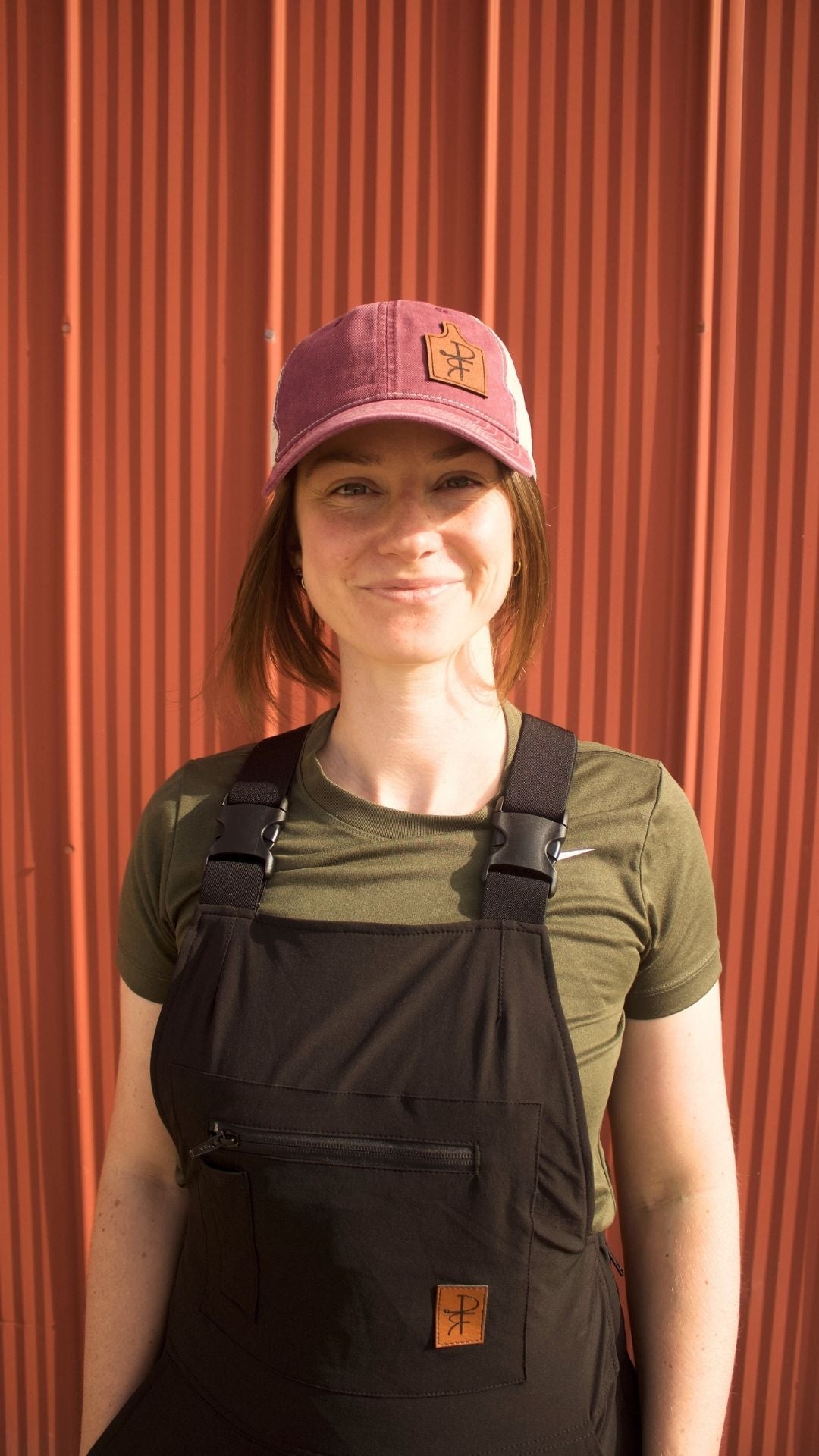 Person wearing a green shirt, black overalls, and a maroon cap against a red metal wall.