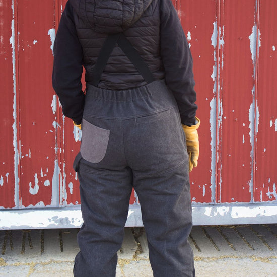 Person wearing a black puffer jacket, black overalls, and a brown beanie standing in front of a red wall.