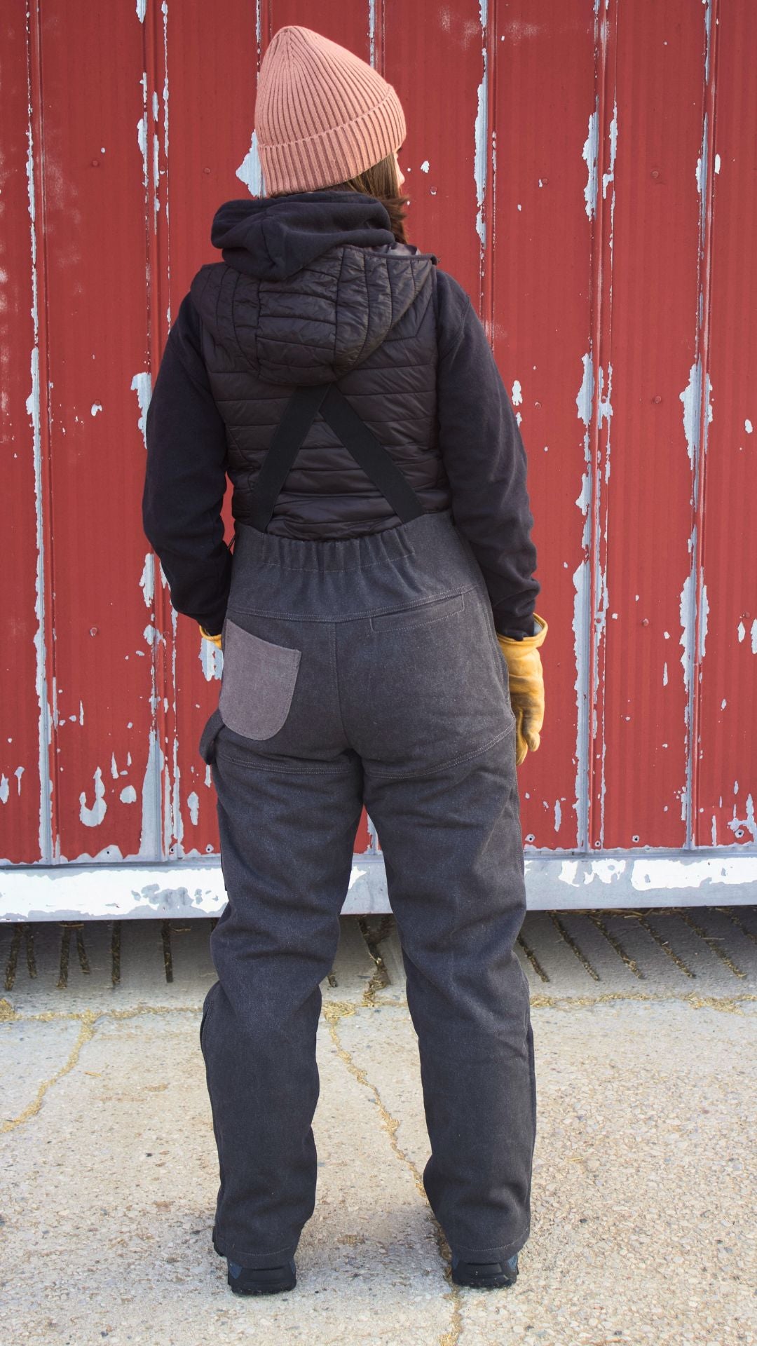 Person wearing a black puffer jacket, black overalls, and a brown beanie standing in front of a red wall.
