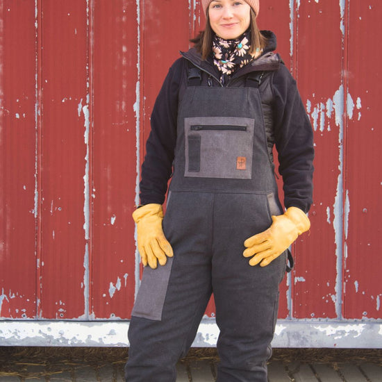 Person wearing a black overall, gloves, and a beanie in front of a red barn.