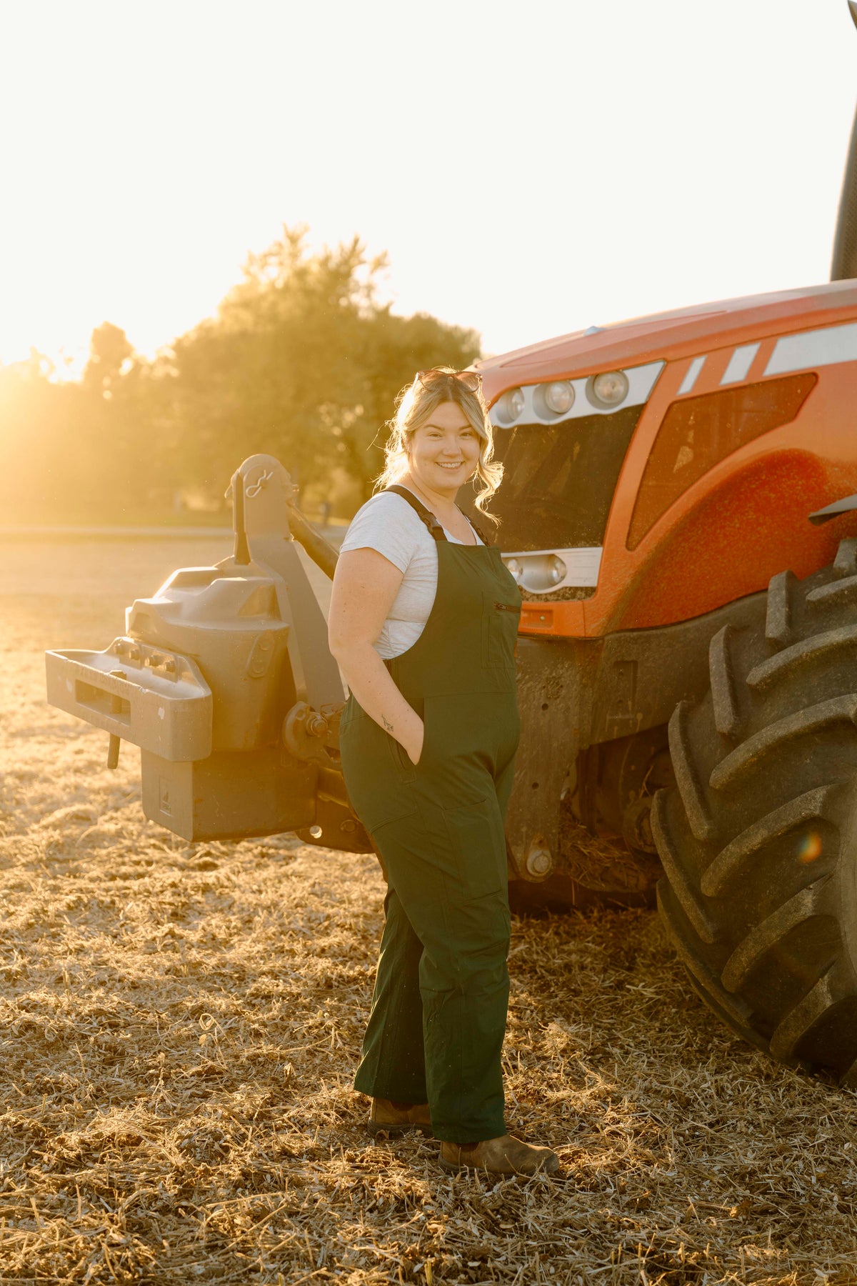 Woman wearing green overalls standing next to a large orange tractor in a field