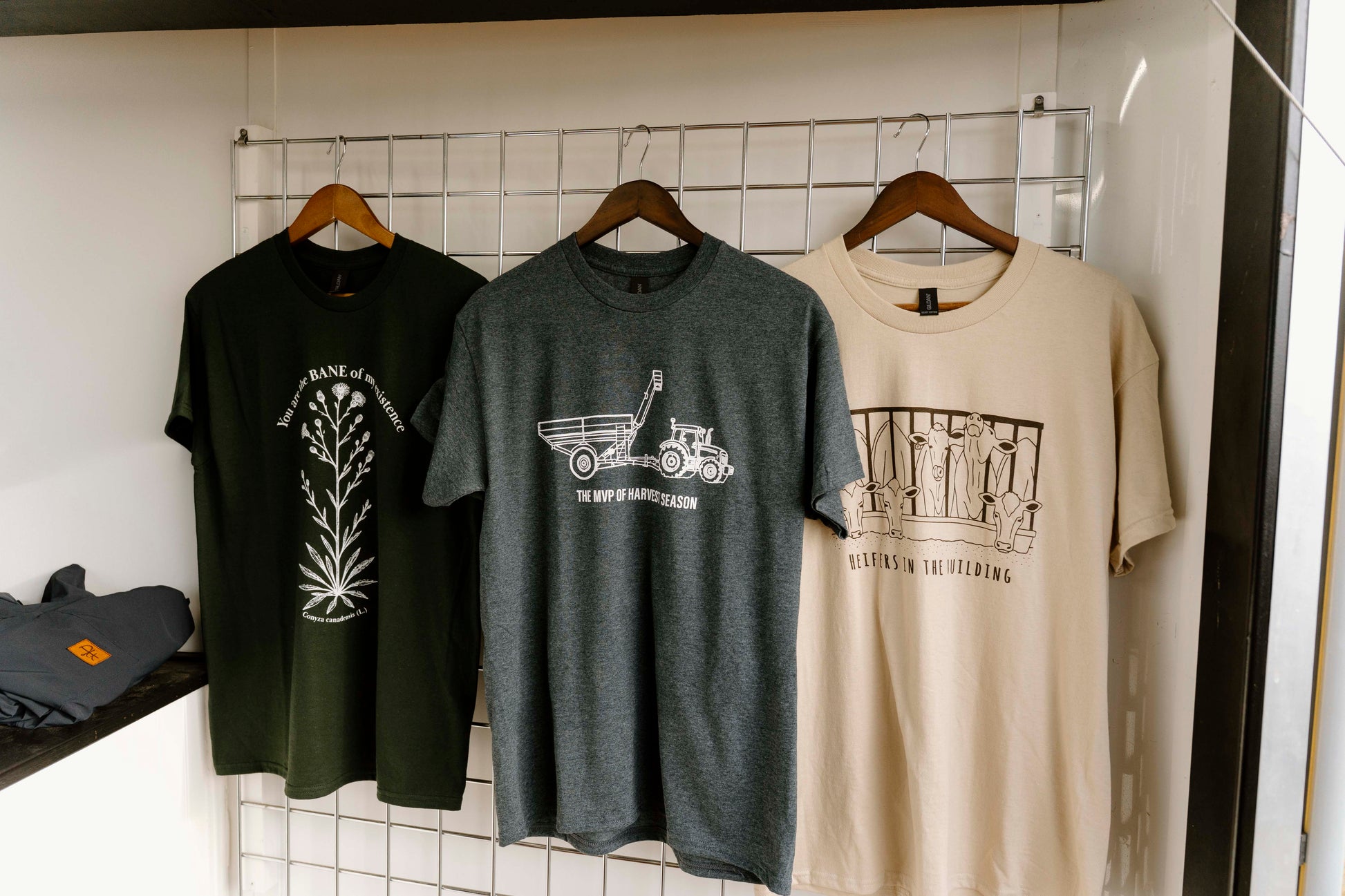 Three t-shirts with different designs hanging on a rack against a white tiled wall.