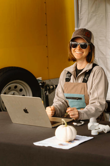 A woman wearing a brown cap, sunglasses, and a beige hoodie sits behind a table under a canopy tent, holding a teal item. A laptop is open on the table, alongside a small white pumpkin, a price list sign, and a black backdrop with a yellow side panel.