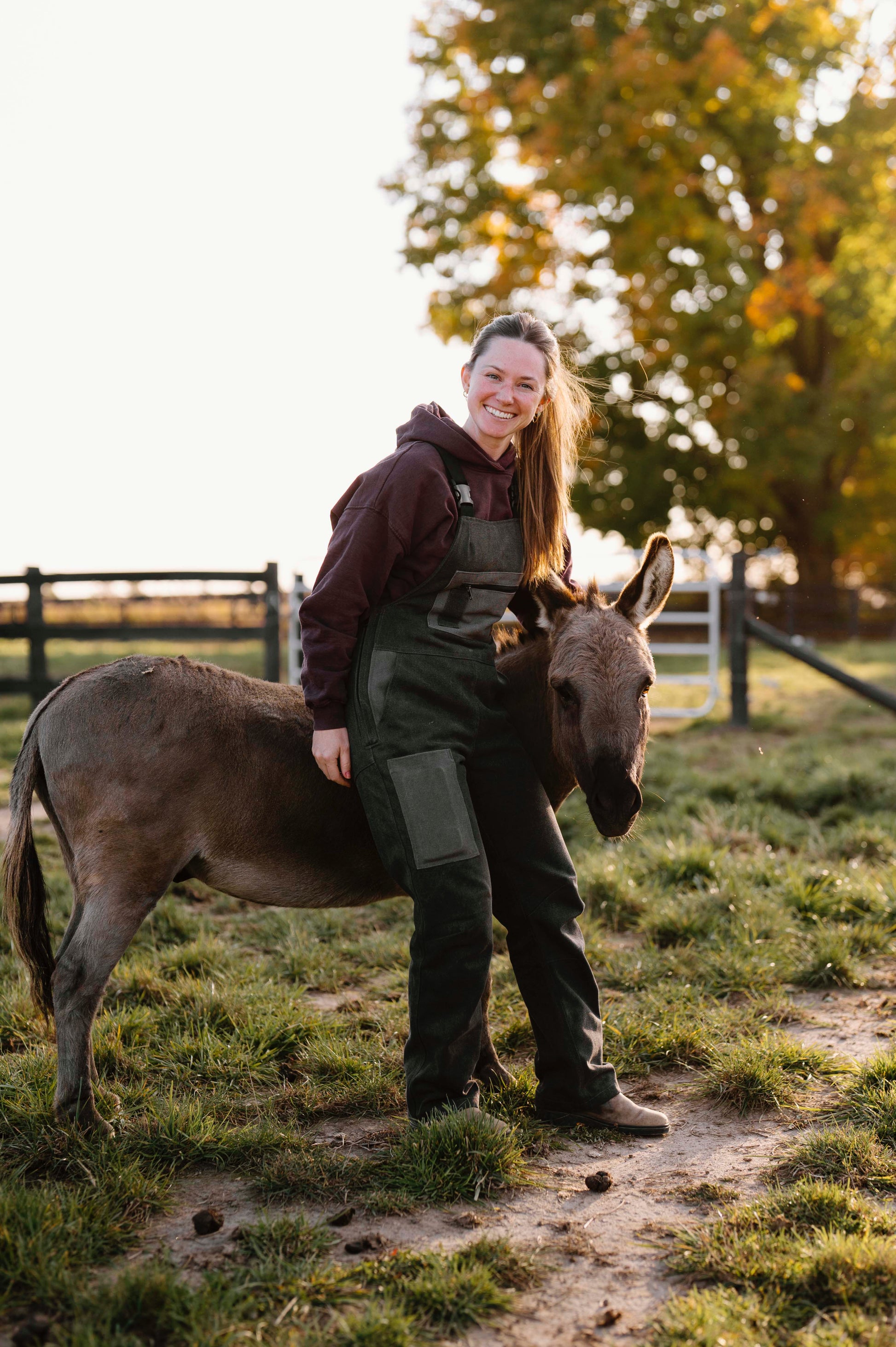 Woman standing next to a donkey wearing grey overalls in a grassy field with trees in the background