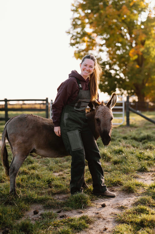 Woman standing next to a donkey wearing grey overalls in a grassy field with trees in the background