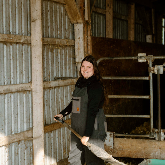 Person holding a broom inside a barn with wooden walls and hay on the floor.