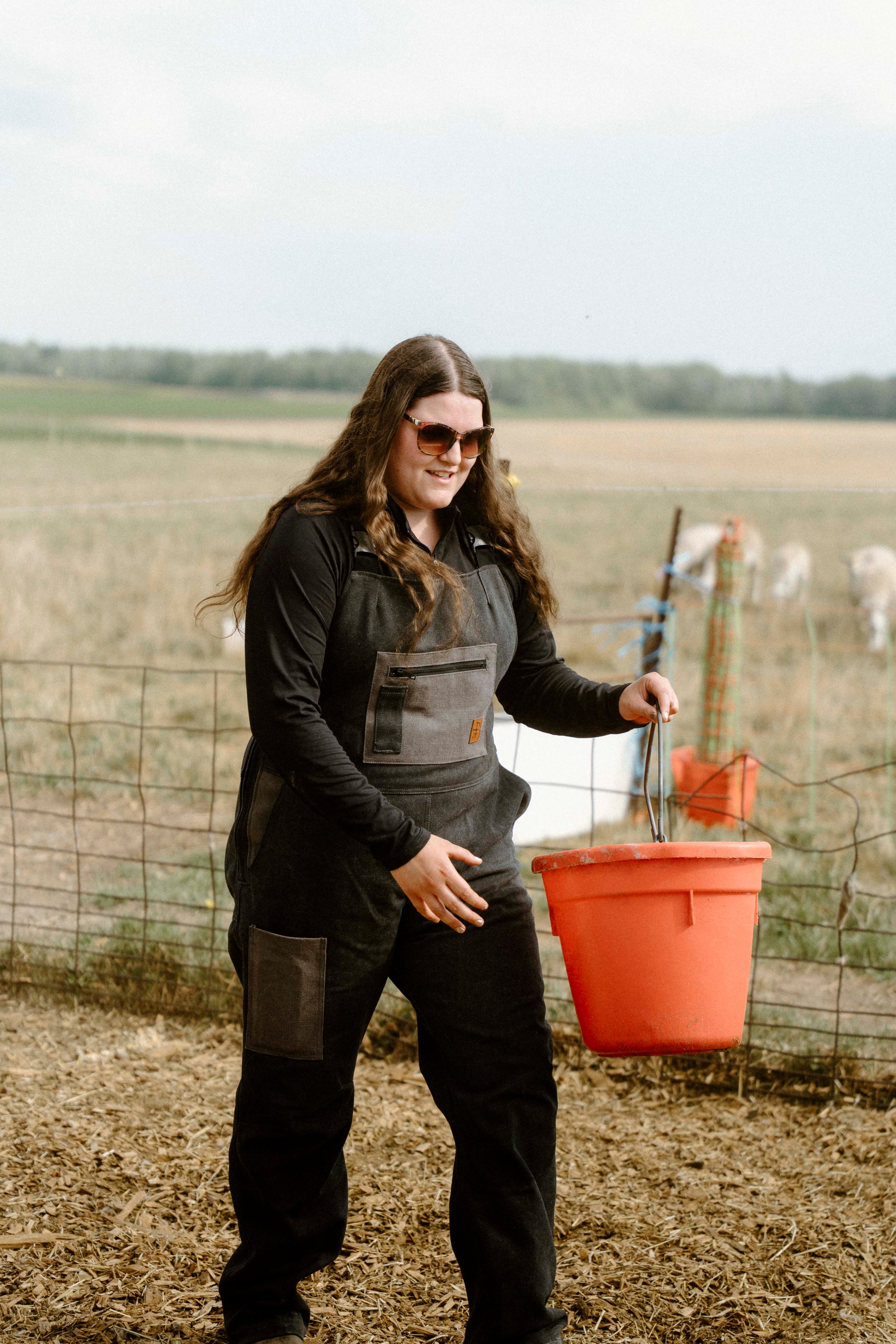 Person standing outdoors with a field and fence in the background wearing dark overalls