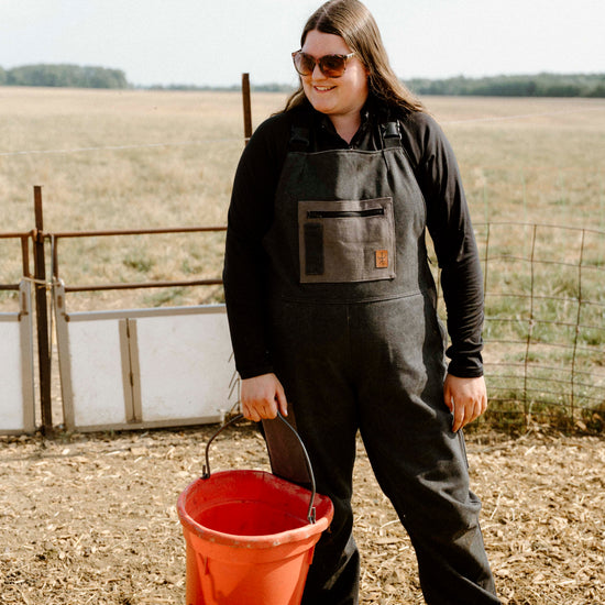 Person wearing a dark overalls and holding a red bucket in an outdoor setting with a field and fence in the background.