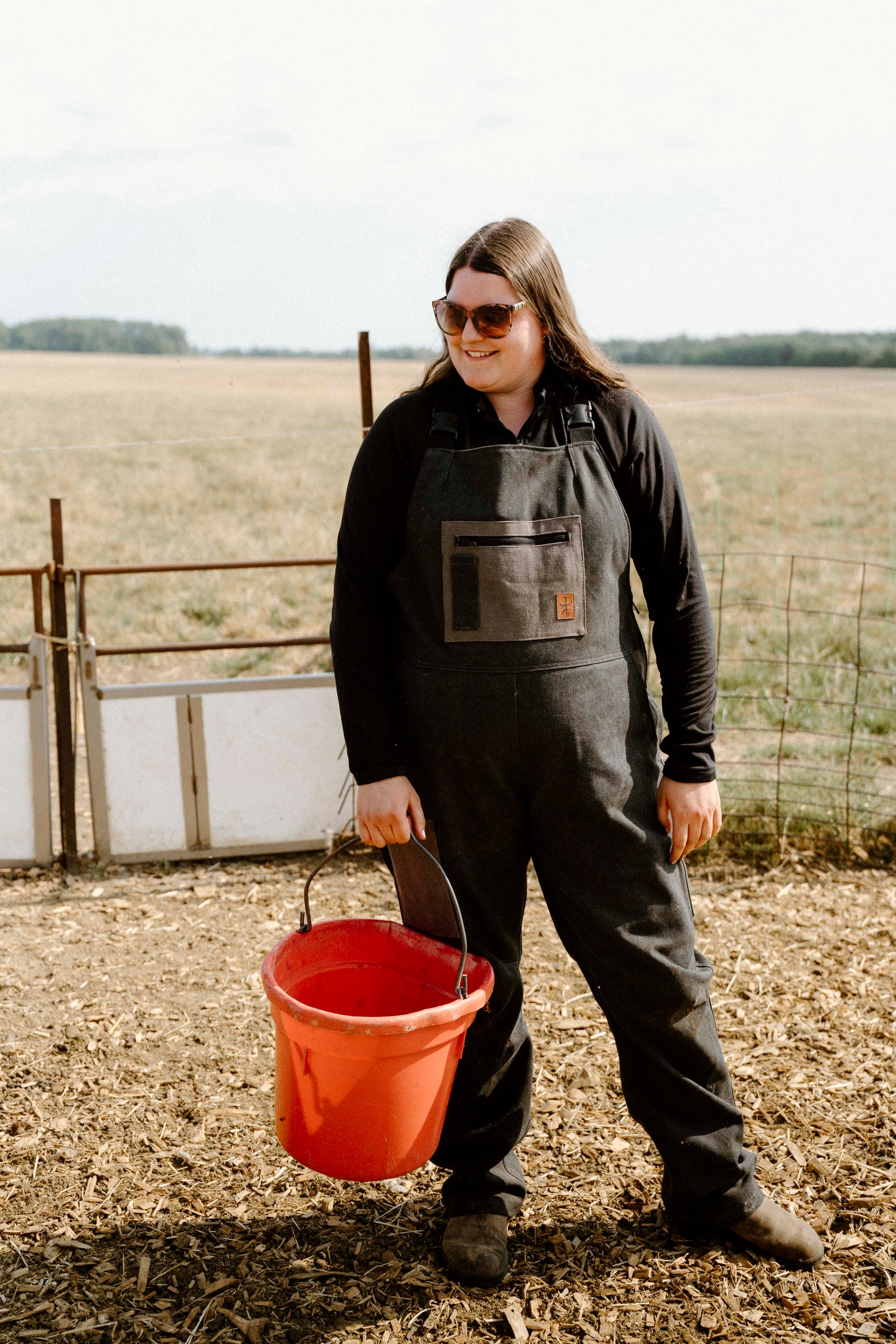 Person wearing a dark overalls and holding a red bucket in an outdoor setting with a field and fence in the background.