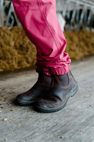 Person wearing pink overalls and black boots on a wooden floor with hay in the background