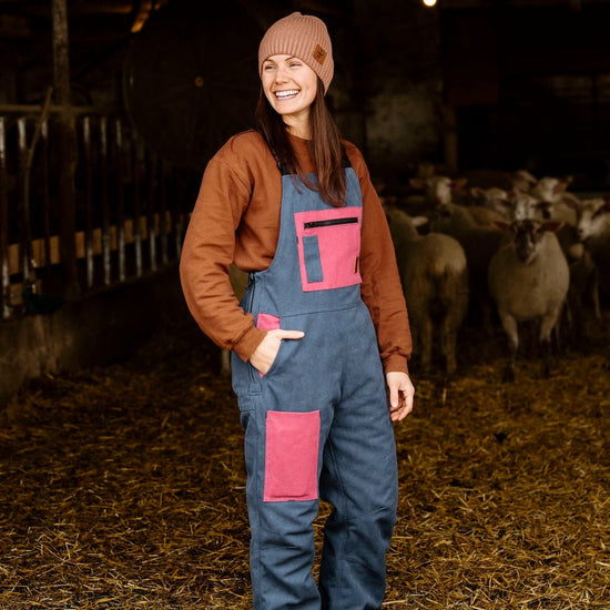 Woman in a barn with sheep, wearing a brown jacket and blue overalls with pink patches.