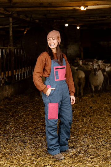 Woman in a barn with sheep, wearing a brown jacket and blue overalls with pink patches.