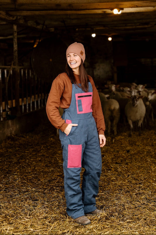 Woman in a barn with sheep, wearing a brown jacket and blue overalls with pink patches.
