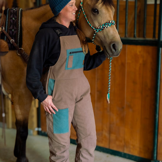A person standing next to a horse, wearing beige and turquoise insulated overalls featuring multiple pockets and a zippered leg.