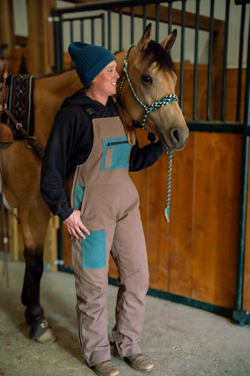A person standing next to a horse, wearing beige and turquoise insulated overalls featuring multiple pockets and a zippered leg.