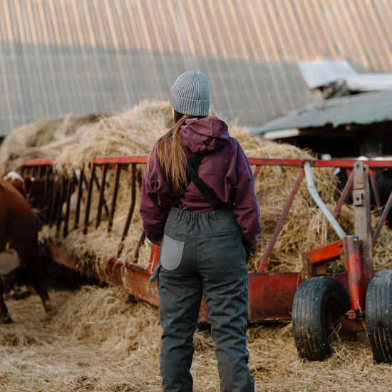 Person wearing dark overalls with grey beanie in-front of hay bale