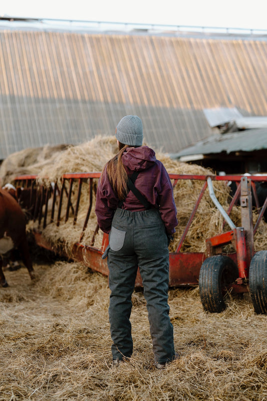 Person wearing dark overalls with grey beanie in-front of hay bale