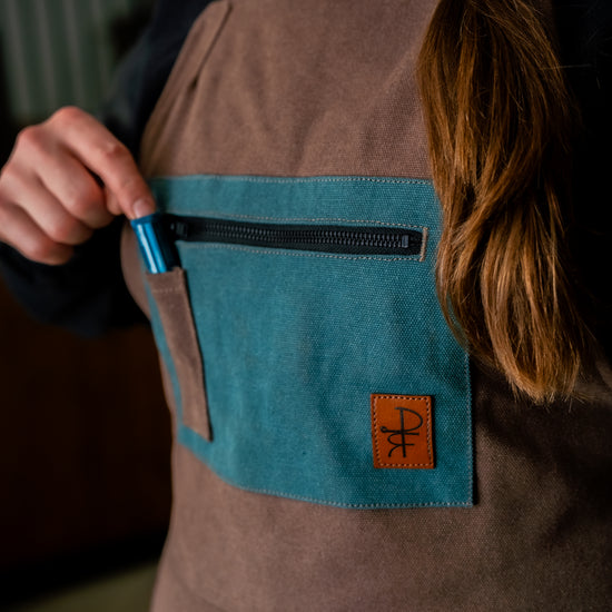 Person wearing brown overalls with teal pockets placing pen into pocket 