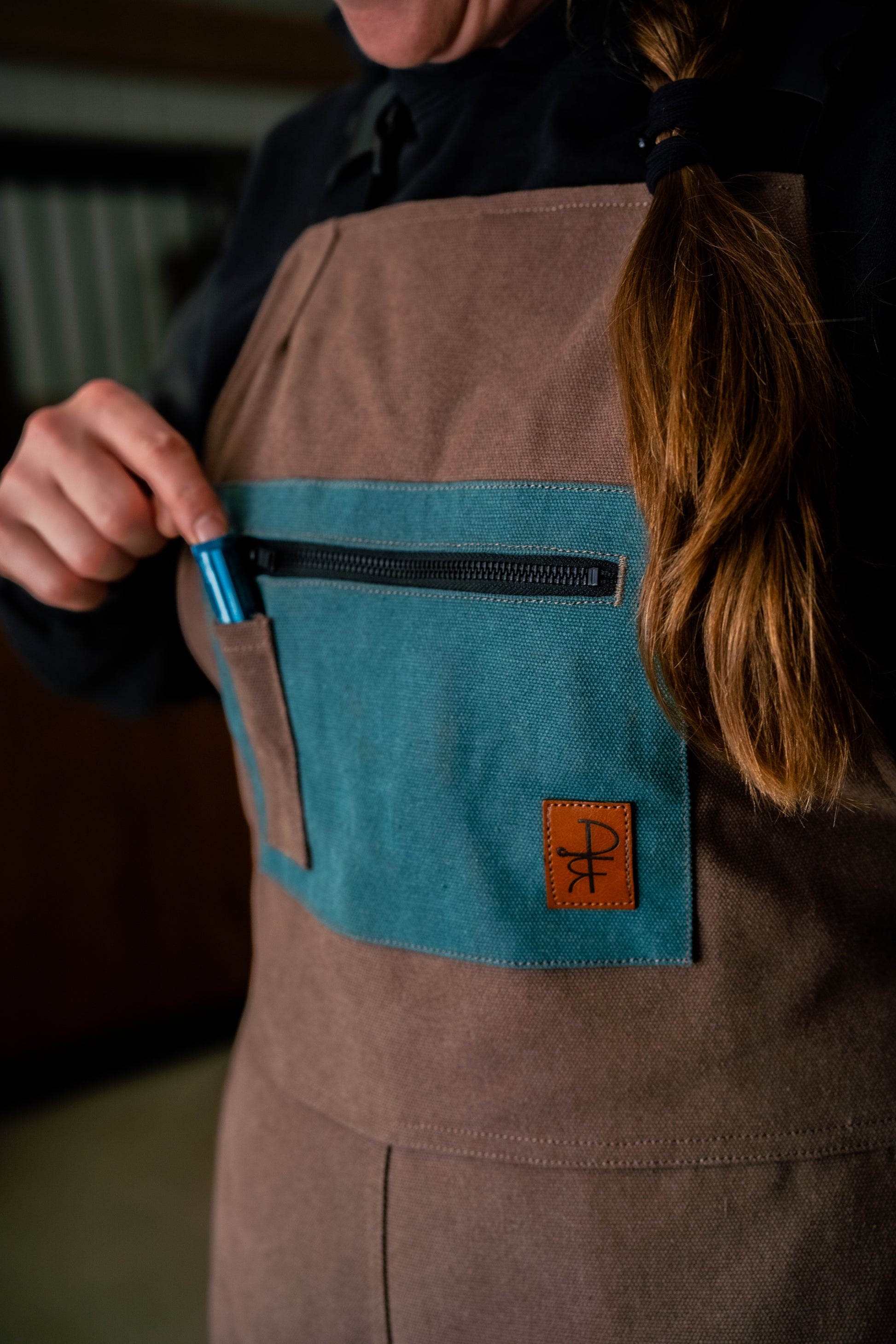 Person wearing brown overalls with teal pockets placing pen into pocket 