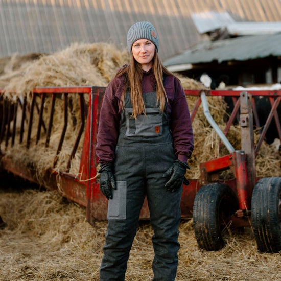 A person standing in front of a hay bale wearing dark charcoal insulated overall with zippered leg, in a farm setting.