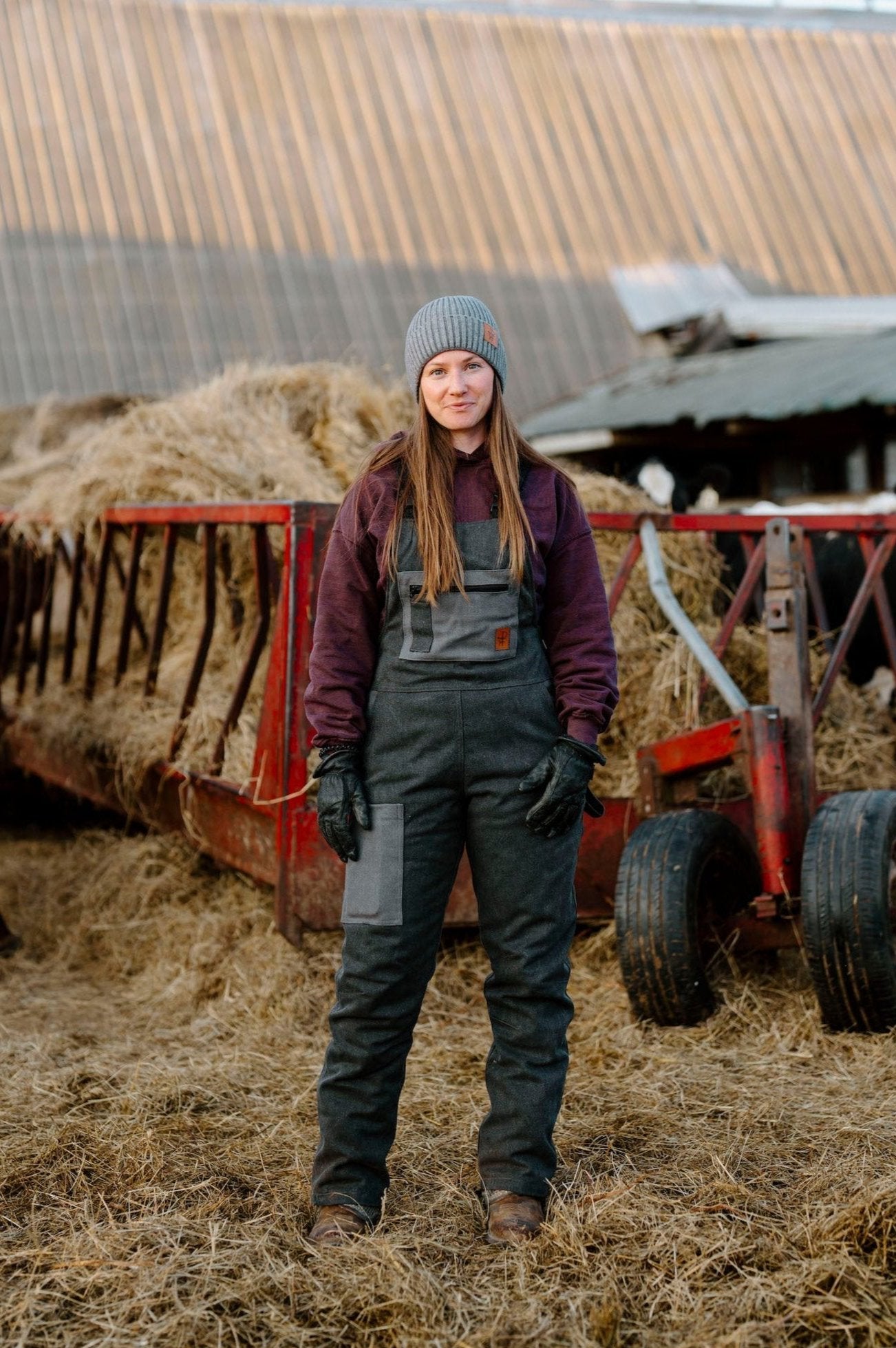 A person standing in front of a hay bale wearing dark charcoal insulated overall with zippered leg, in a farm setting.