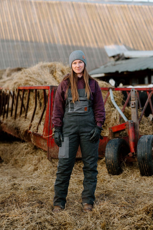 A person standing in front of a hay bale wearing dark charcoal insulated overall with zippered leg, in a farm setting.