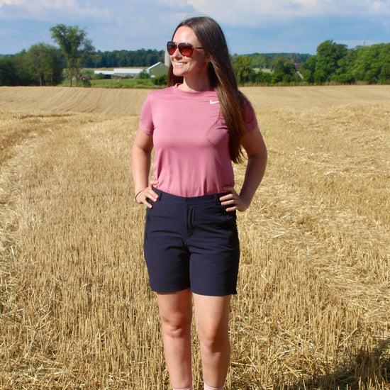 Person standing in sunny field with farm in background. wearing navy shorts, pink shirt and brown boots 