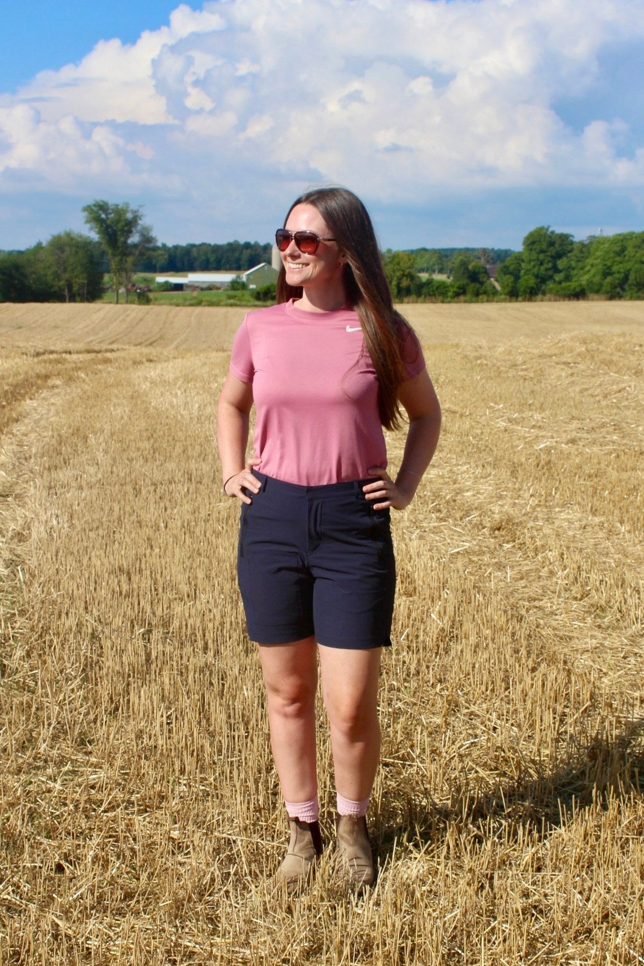 Person standing in sunny field with farm in background. wearing navy shorts, pink shirt and brown boots 